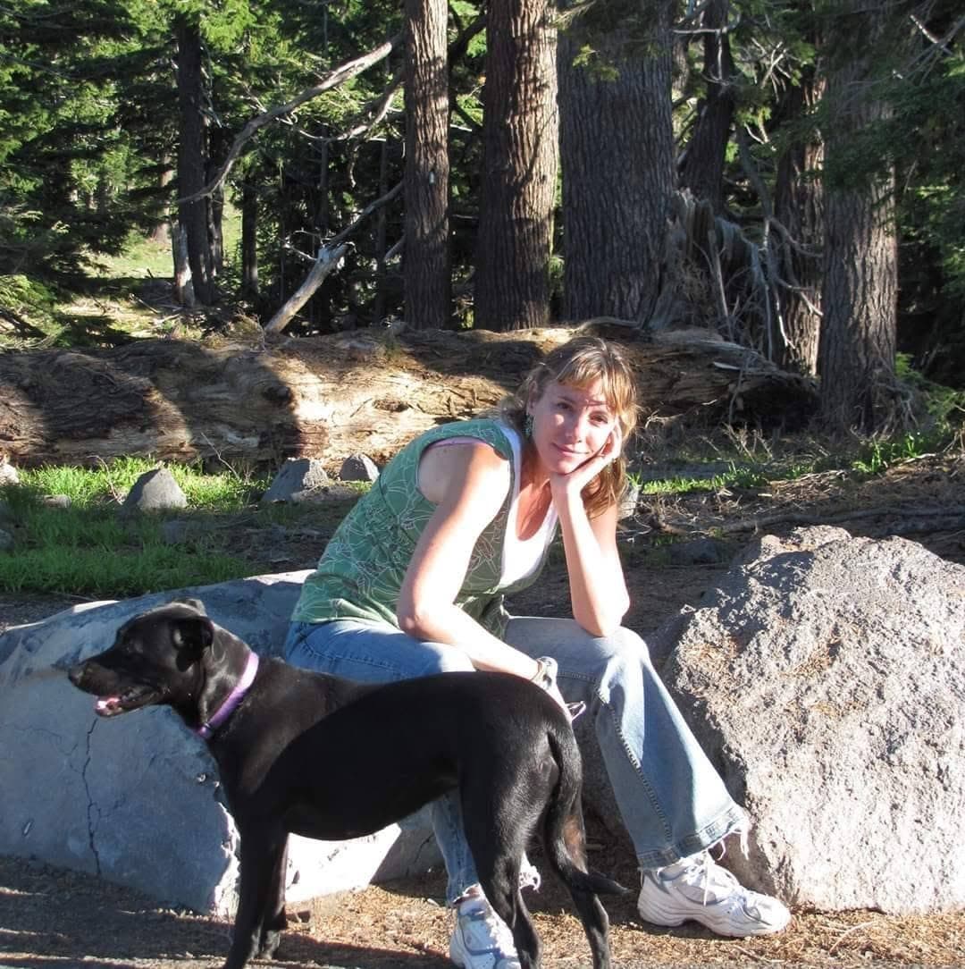 Elsa C White sitting on a boulder in a forest clearing with her dog.